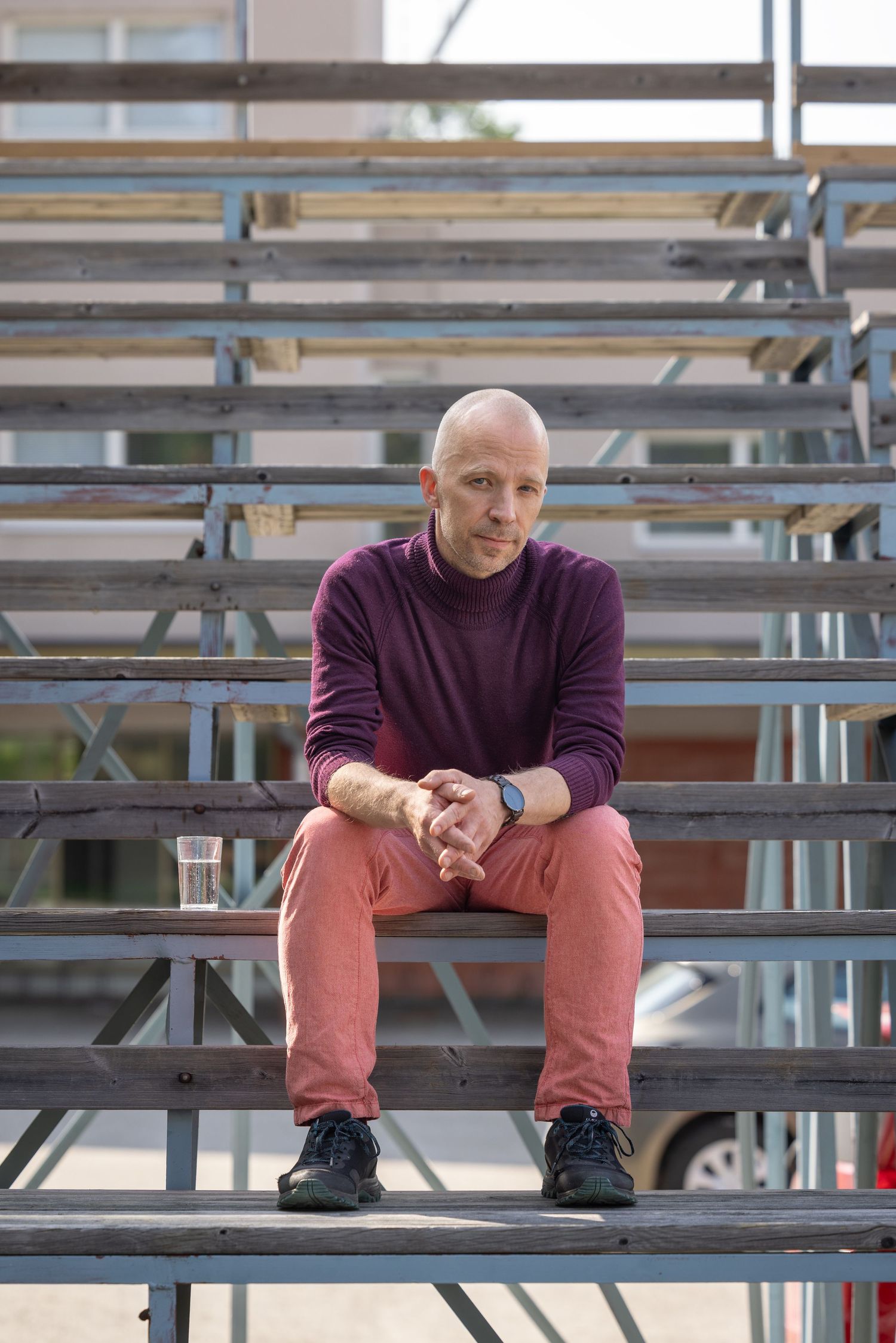 Man wearing purple sweater and salmon colored pants sits casually on outdoor metal bleachers.