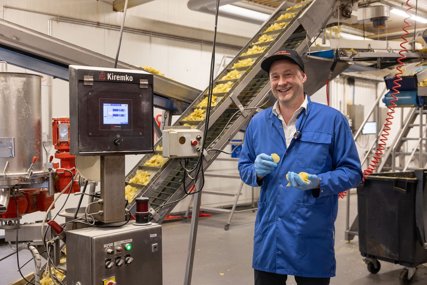 Man in blue lab coat and hat smiling in a food processing facility next to industrial machinery.