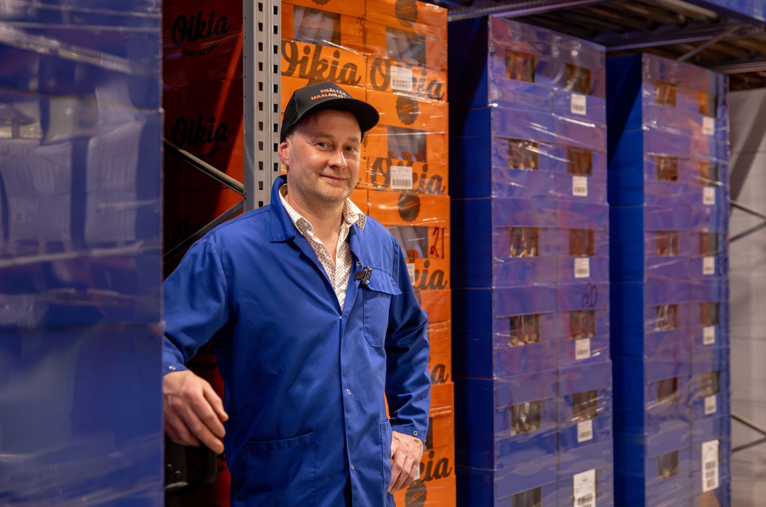 Man in blue work uniform smiling in warehouse storage facility with stacked orange branded crates.