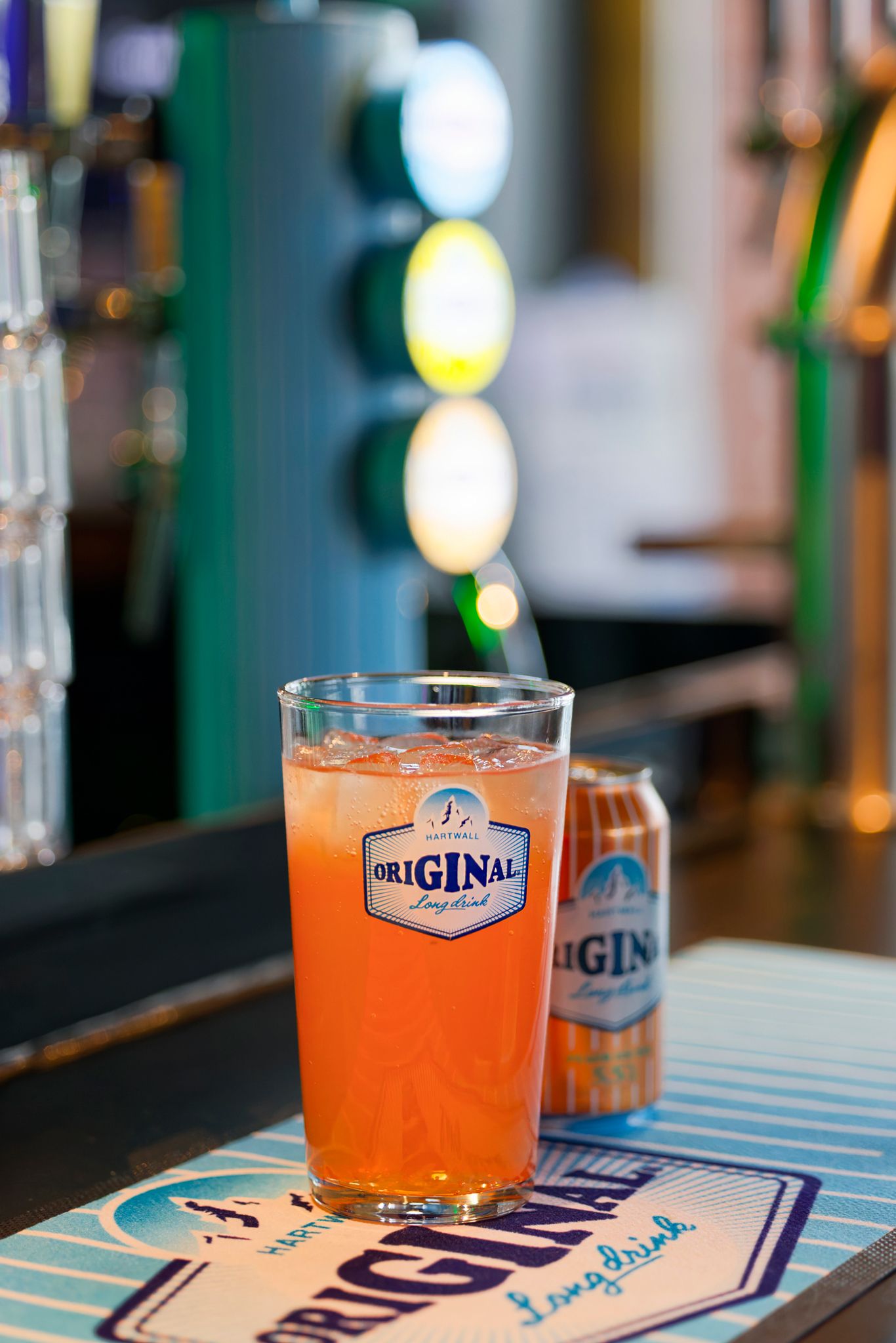 A glass of Original Long Drink cocktail on a bar counter with a can and tap handles in the background.