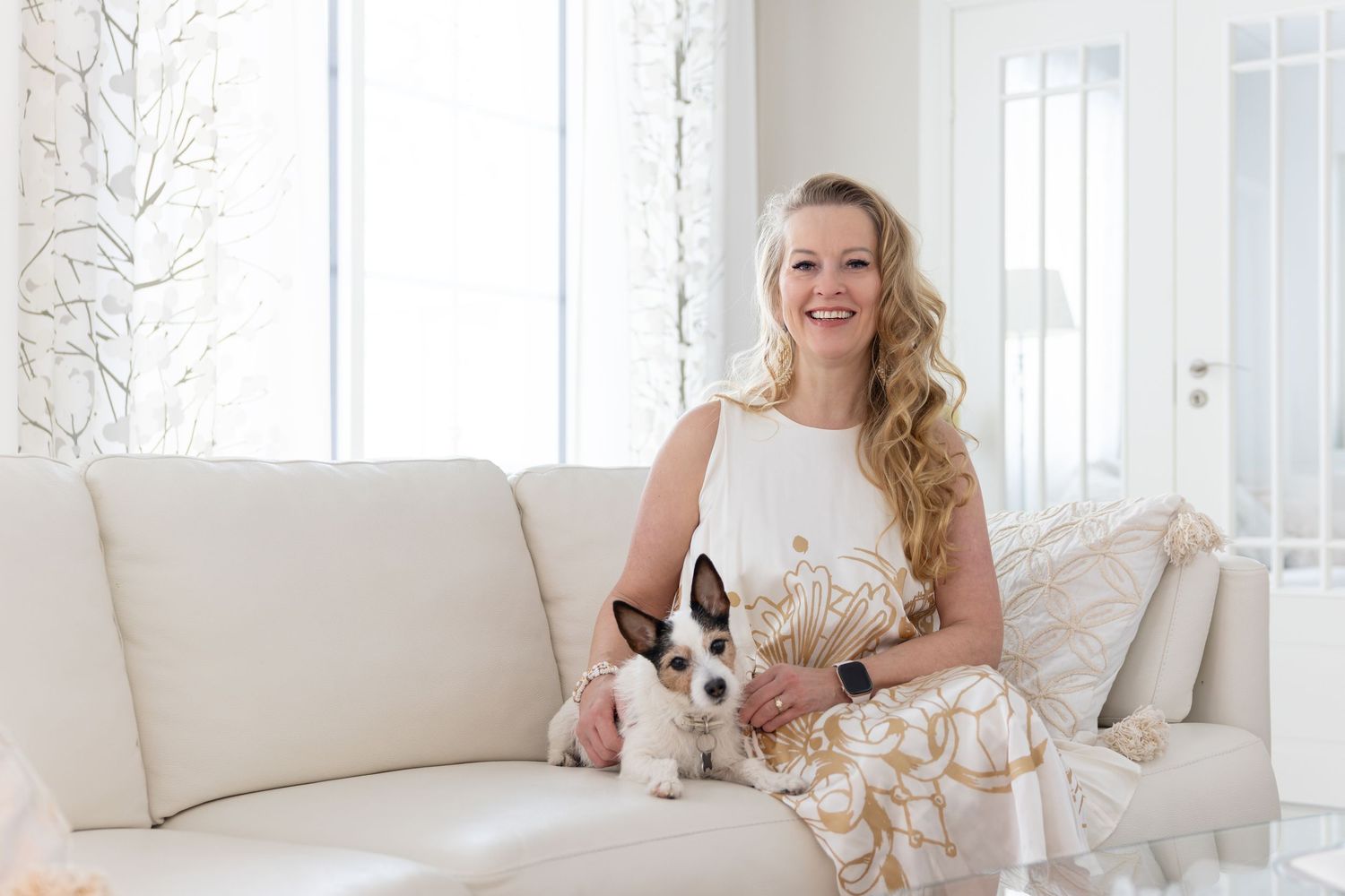Woman in white dress smiling on cream sofa with small Jack Russell terrier dog in bright white living room.