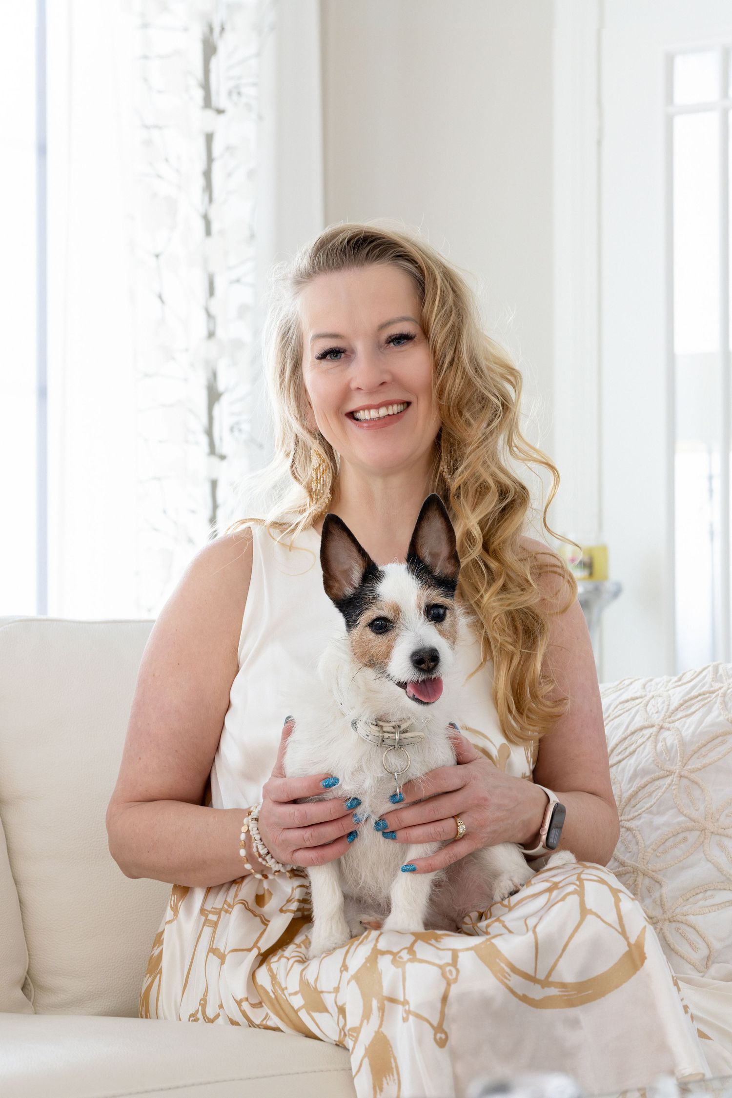 Smiling blonde woman with blue nails holding a small Jack Russell terrier dog while sitting on a white sofa.