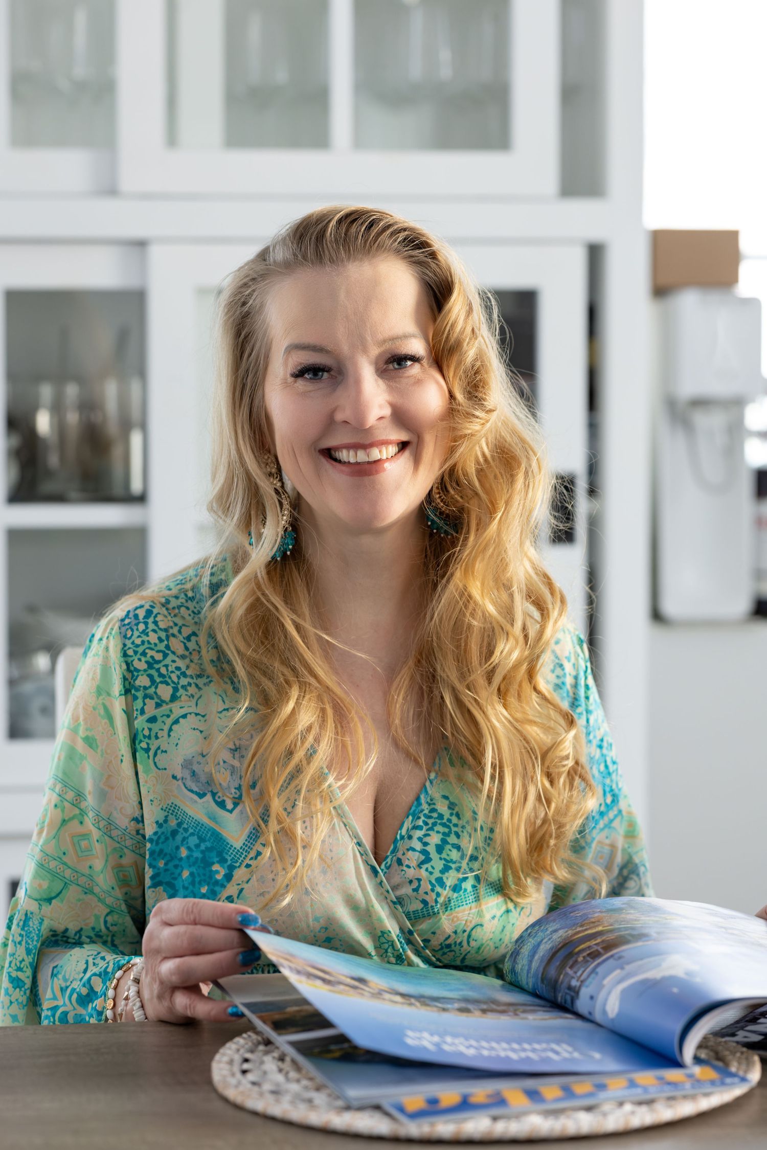 Smiling blonde woman in turquoise floral blouse reading a magazine at a table in a bright white kitchen.