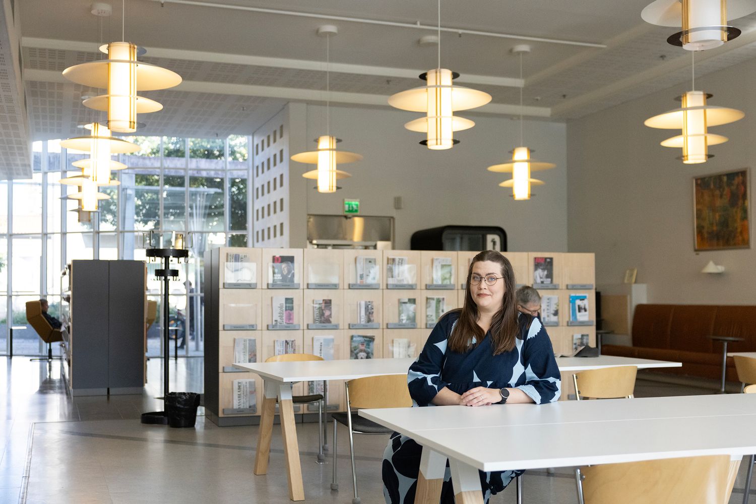 Unikkomekkoon pukeutunut nainen istuu kirjaston lukusalissa, katossa designvalaisimia.