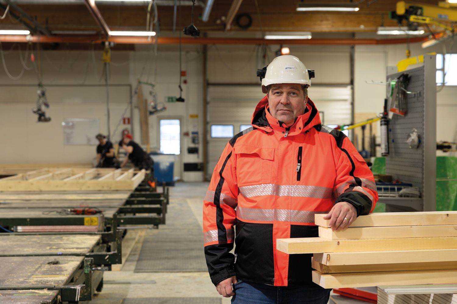 Kypärään ja suojatakkiin pukeutunut mies seisoo puuntyöstöhallissa talotehtaalla. Taustalla työntekijöitä ja isoja puisia seinäelementtejä.