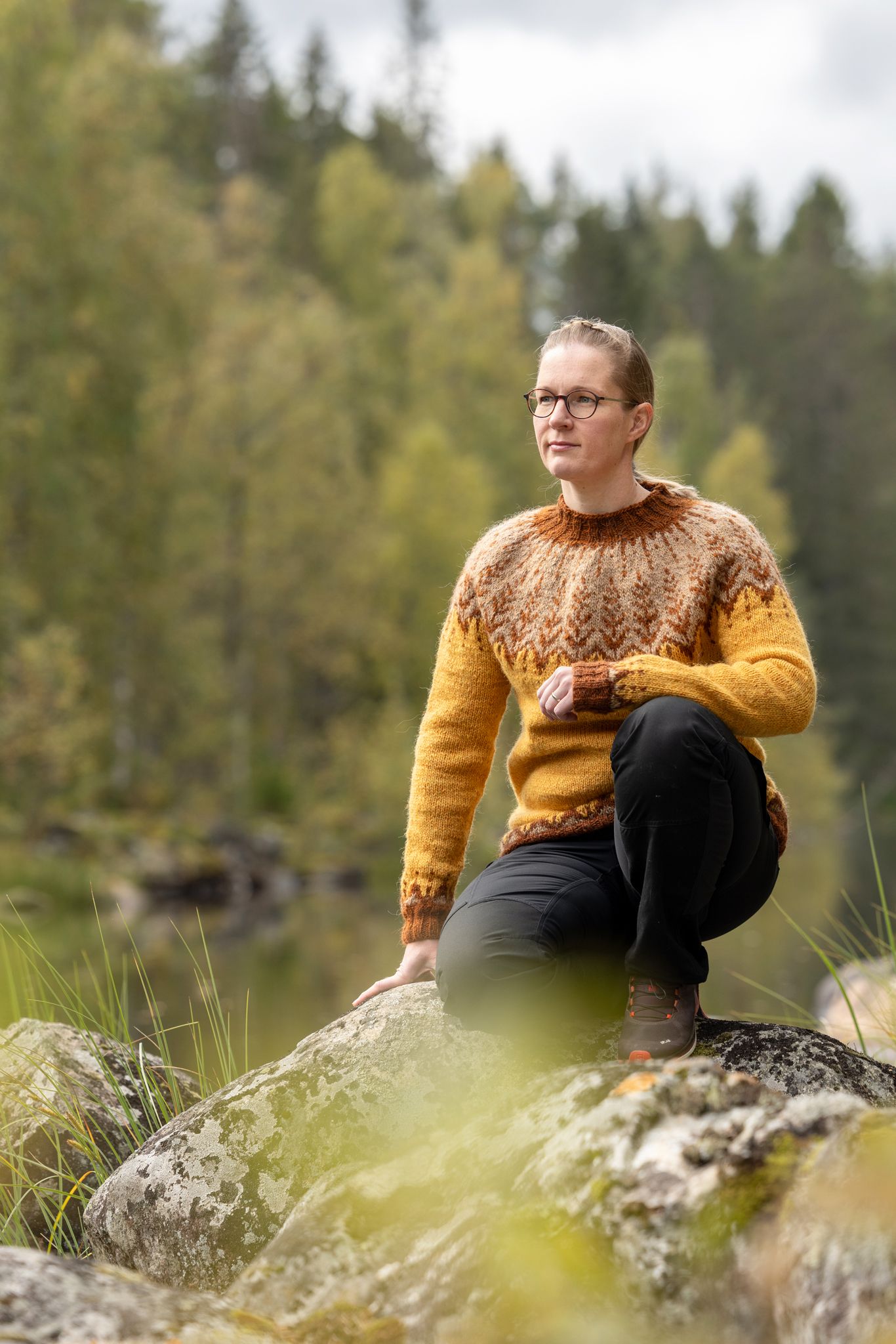 Neulepuseroon pukeutunut nainen istuu kyykyssä rantakivillä.