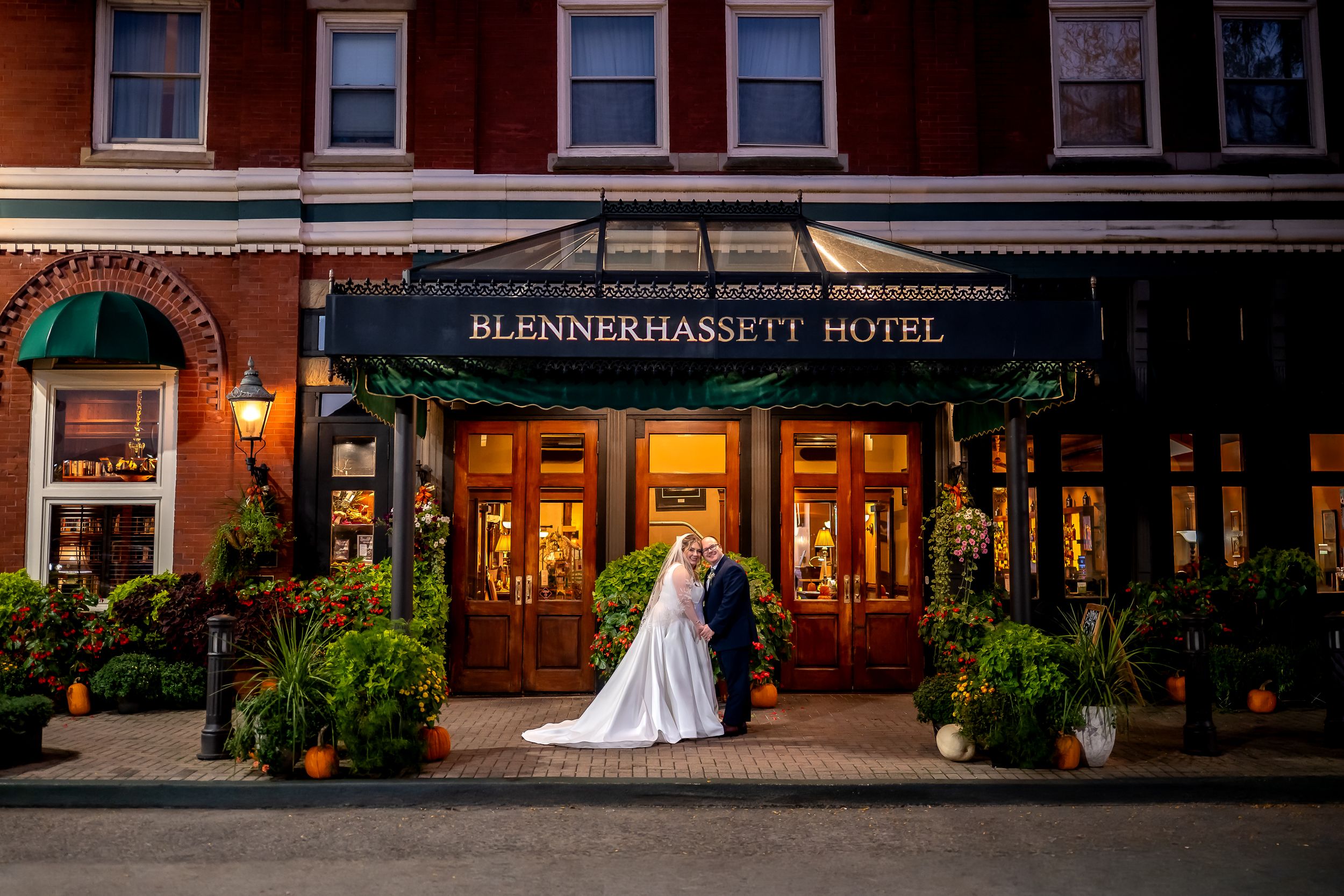 Historic brick buildings with storefronts line a cobblestone street at dusk with warm lighting from windows and lamps.