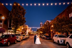 String lights illuminate a charming small town street at dusk with historic buildings and parked cars.