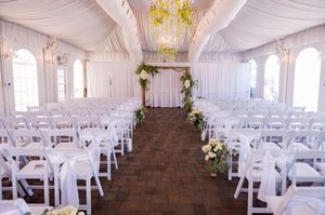 Elegant wedding ceremony setup with white chairs and floral arch in a draped tent venue with chandeliers.