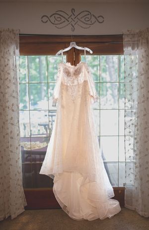 White lace wedding dress hanging in window with sheer curtains and natural light.