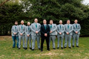 Wedding party members wearing light gray suits and blue ties pose outdoors on a grassy lawn.