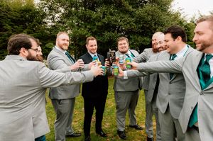 Groomsmen in gray suits holding beverages in a toast formation during an outdoor wedding celebration.