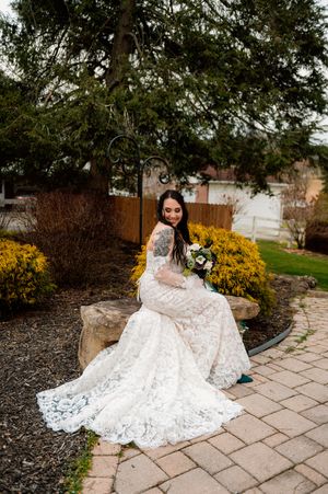 A flowing lace wedding dress spreads elegantly across stone pavers in a garden setting.