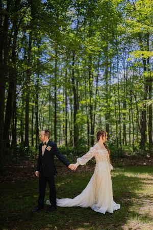 A romantic first look moment captured in a sunlit forest setting with flowing white dress and formal attire.