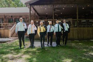 Group of groomsmen dressed in light blue shirts, dark pants, and suspenders walk together on a lawn outside a rustic venue.
