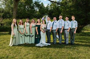 Wedding party poses outdoors in light sage green and white attire against a natural backdrop of trees and grass.