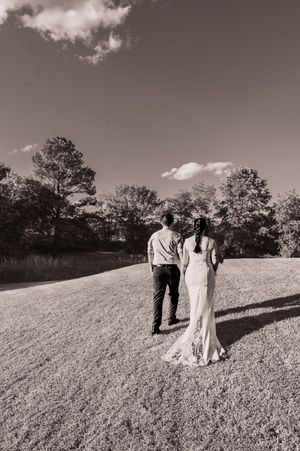 Black and white portrait of newlyweds walking up a grassy hillside under a cloudy sky.