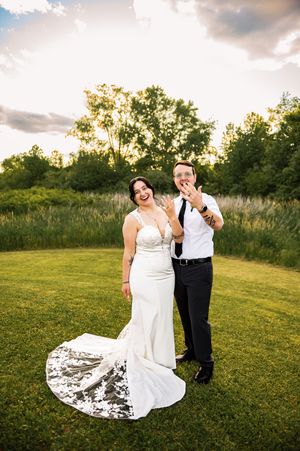 A newlywed couple poses playfully on a grass lawn at sunset with a flowing white wedding dress train.