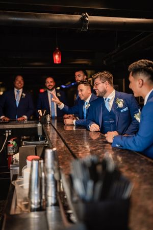 Wedding party in blue suits gathered around dark wooden bar for casual photos.