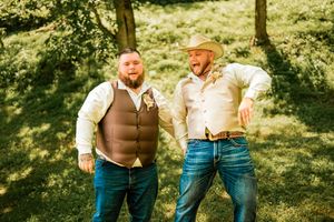 Two men in casual western attire laughing together in a sunny outdoor setting.