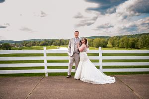 Wedding couple standing together against white fence with rolling hills in background.