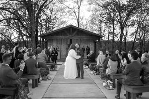 Wedding ceremony takes place outdoors with guests seated on wooden benches in front of a rustic pavilion.