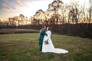 A couple walks together in a field at sunset with trees silhouetted against a pink and orange sky.