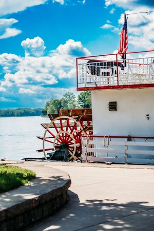 A red and white riverboat paddle wheel against a bright blue sky with fluffy clouds.