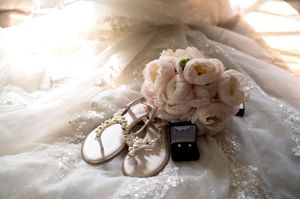 Wedding accessories including ballet flats, a ring box and pink peonies arranged on lace fabric.