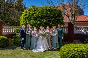 Wedding party poses in a garden setting with red brick building and manicured hedges on a sunny day.