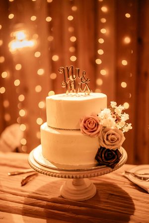 An elegant two-tier white wedding cake with rose decorations and Mr & Mrs topper against twinkle lights.
