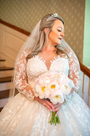 A bride in an elegant lace wedding gown holds a white floral bouquet while posing near a staircase.