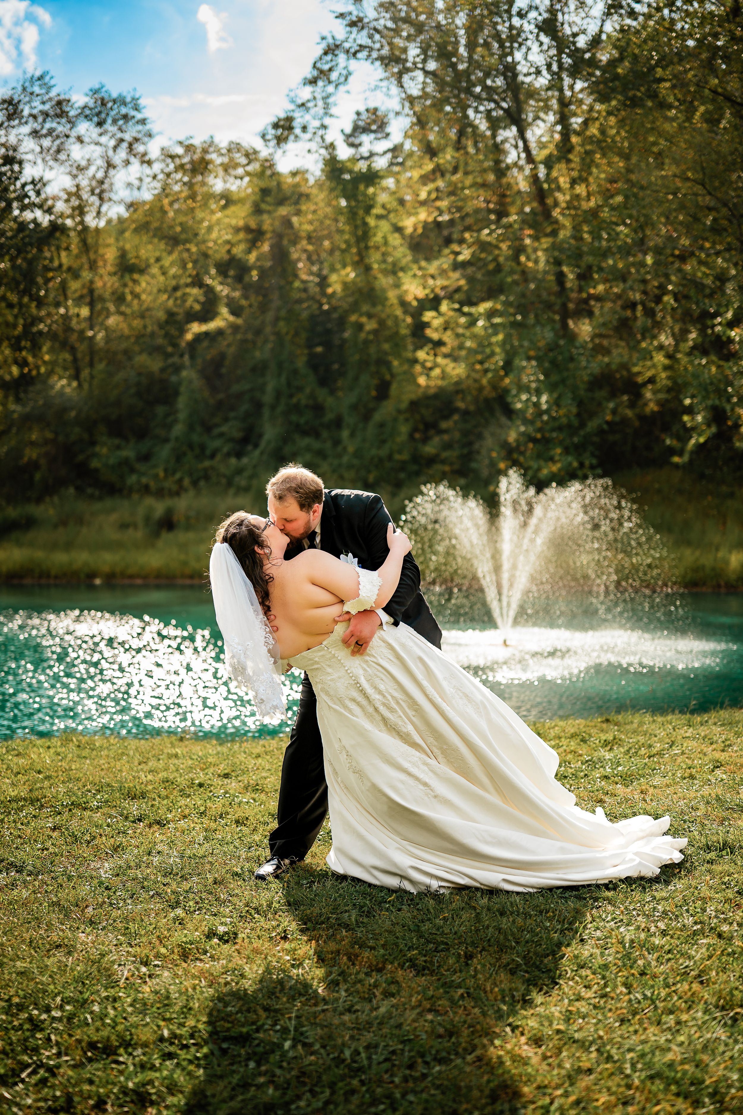 A romantic wedding couple shares a dip kiss on grass with a fountain spraying in the background on a sunny day.
