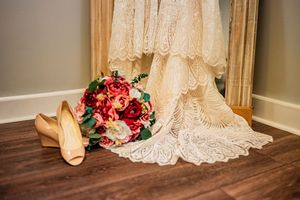 Wedding details with ivory lace dress, beige heels, and vibrant red and pink bridal bouquet on wooden floor.