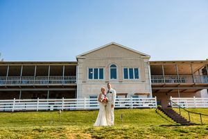 A couple stands on a grassy hill in front of a white colonial-style building with wrap-around porches.