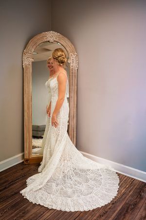 A wedding dress with intricate lace details and a dramatic train is showcased against dark hardwood floors and gray walls.