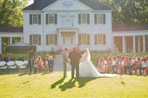 A wedding ceremony takes place in front of a grand white colonial mansion at sunset.