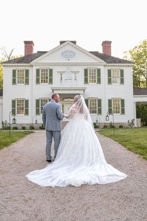 A couple in wedding attire stands on a gravel path facing a grand white colonial house.