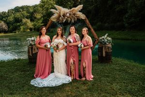 Wedding party in coral pink bridesmaid dresses pose together on grassy lawn near lake during outdoor celebration.