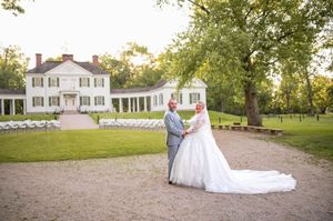 A wedding couple embraces on the grounds of an elegant white colonial estate with curved wings.