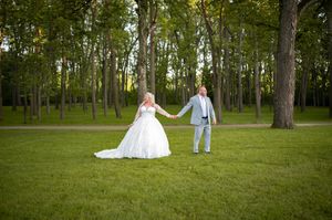 A couple in wedding attire holds hands while walking through a lush green forest.