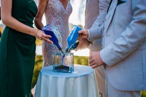 Two people in formal attire stand together during a wedding ceremony with decorative floral arrangements in the background.