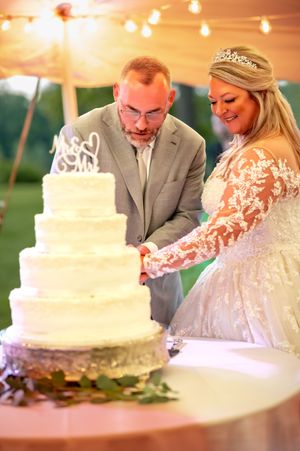 Couple cutting their elegant white tiered wedding cake under warm tent lighting.