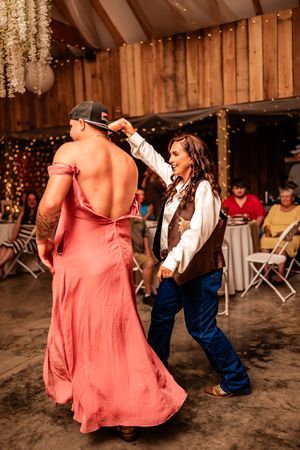 Pink-dressed dancers twirl and dance in a rustic barn reception with string lights and wooden beams.