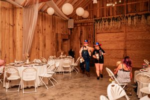 Wedding reception setup inside a rustic wooden barn with white chairs and round tables under hanging globe lights.