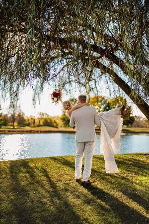Couple in white attire walking together under willow trees by a tranquil lake at sunset.