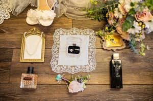 Wedding details arranged on wooden surface with perfume bottles, frames and floral accents.