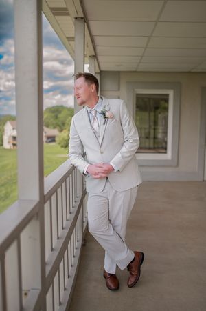 Person in a light gray suit and boutonniere leans against a railing on a covered porch.