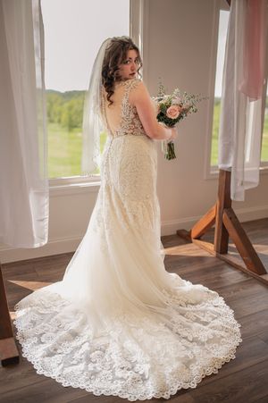 A bride in a stunning lace wedding gown with a flowing train poses near sunlit windows in an elegant venue.