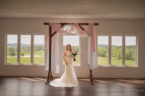 Bride in fitted lace gown poses in sunlit room with large windows and draped fabric decor.
